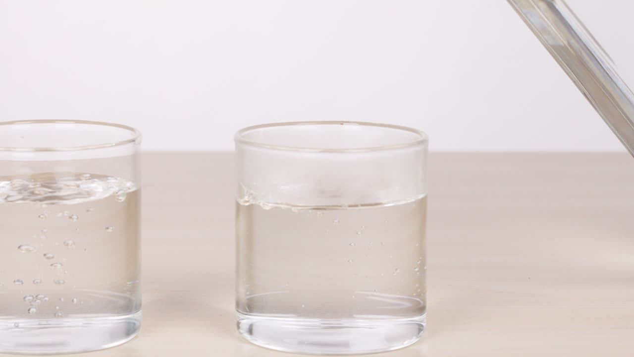 Clear water poured from bottle into two empty glasses on table, bright even lighting