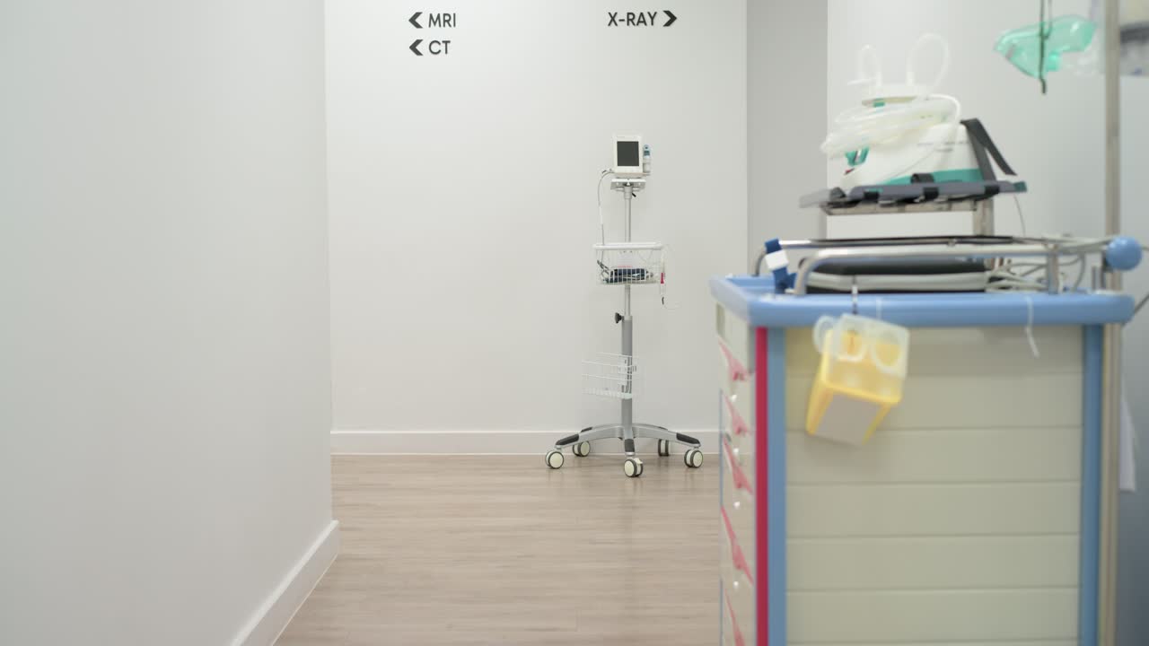 Um carrinho de suprimentos com rodas está entrando pela direita e estacionando para realizar exames de imagem perto do monitor de sinais vitais, próximo às placas de radiologia.