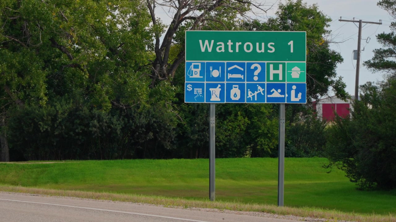 Static close-up telephoto shot of a highway sign outside Watrous, Saskatchewan, with trees behind and roadway visible in the foreground