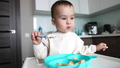 dinner time for a cute little kid. baby sits at feeding table eating from spoon. close up. blurred backdrop.