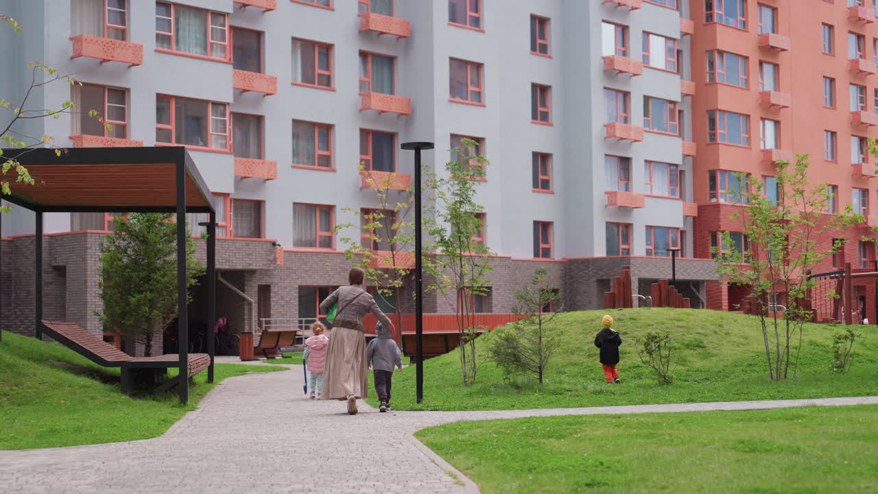 Teacher walks with group of young children through green residential courtyard surrounded by colourful apartment buildings, guiding them safely during outdoor activity under cloudy sky