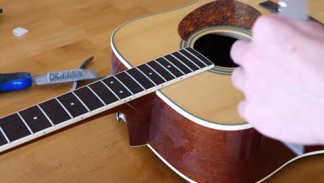 close up hands of a luthier craftsman measuring and leveling an acoustic guitar neck fretboard on a wood workshop bench with lutherie tools slide left