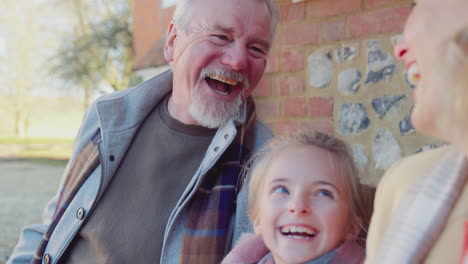 Grandparents-With-Granddaughter-Outside-House-Sitting-On-Seat-Getting-Ready-To-Go-For-Winter-Walk