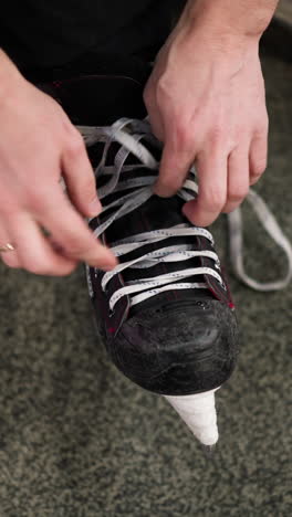 vista de primer plano de las manos soltando los cordones de un patín de hielo negro con acentos rojos, en un piso alfombrado gris, la persona está usando pantalones negros