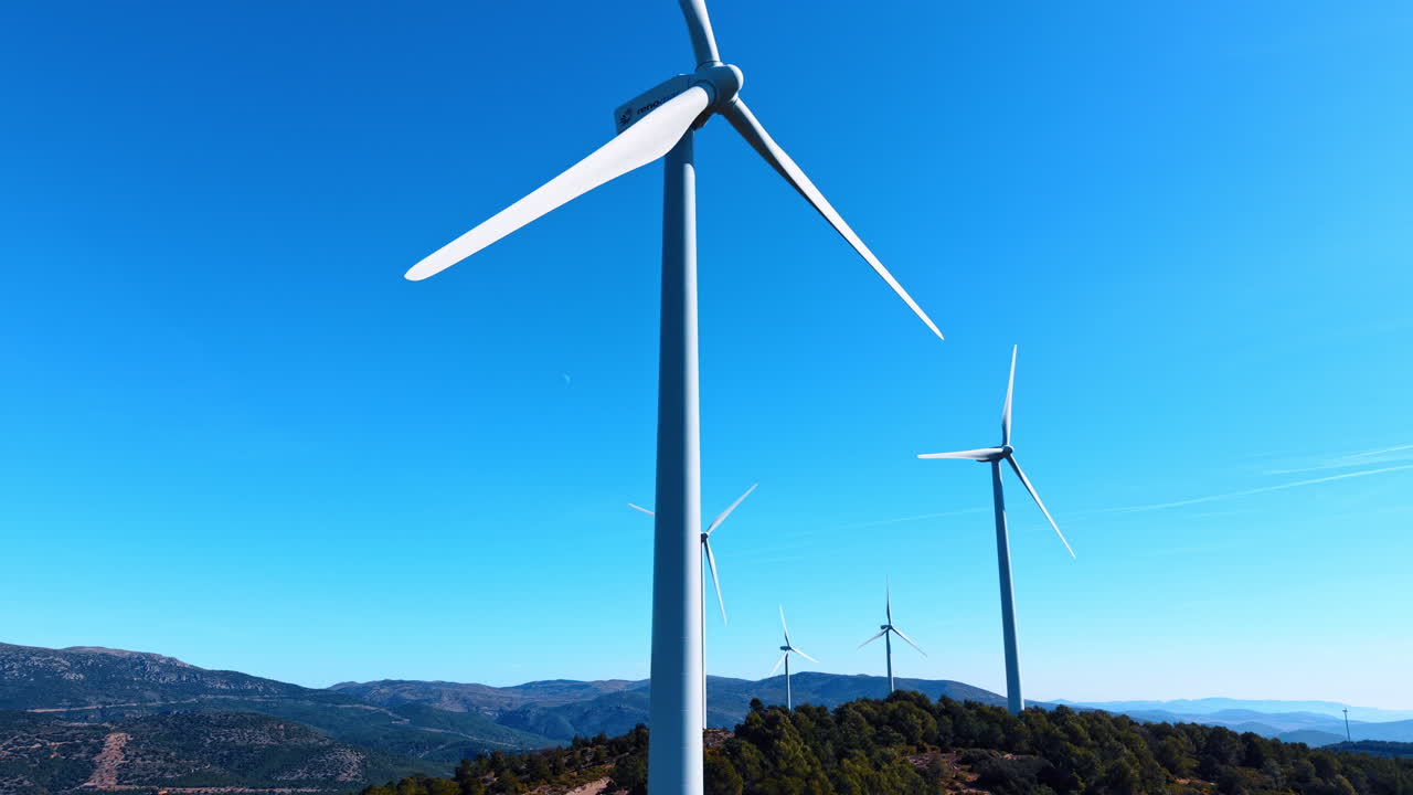 Alicante, Spagna, 8 agosto 2025: Turbine eoliche bianche su crinale montuoso spagnolo. Vista aerea dal basso di turbine eoliche bianche su montagne spagnole boscose sotto un cielo azzurro limpido.