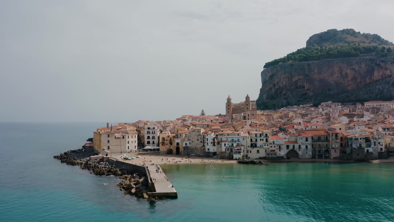 Aerial orbit drone shot along the Cefalu beach. Small historic town near Palermo city by the Mediterranean sea in Sicily. Pier stretching into the sea. Charming Italian village.