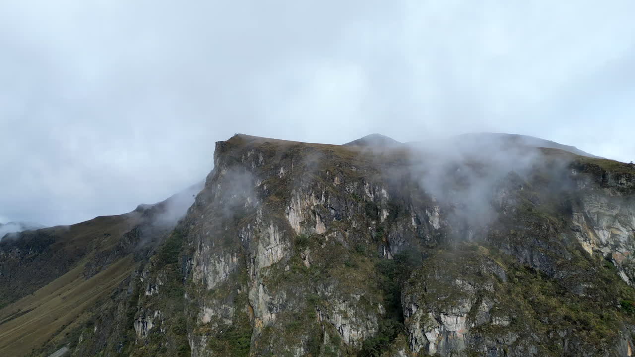 Drone towards a misty mountain in El Cajas, Ecuador. The footage captures rugged stone cliffs, dense tree cover, and light clouds surrounding the peak.