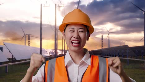 Close Up Of Asian Female Engineer With Safety Helmet Screaming Goal And Dancing Celebrating With Solar Panel and Wind Turbines