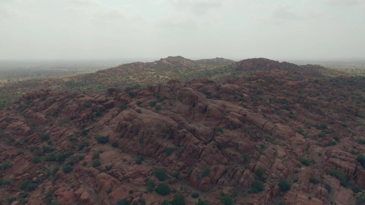 Ampio panorama drone su colline di granito rossastro nel deserto del Thar, Sindh, Pakistan, con creste arrotondate che digradano verso pianure sabbiose e brumose con rada vegetazione