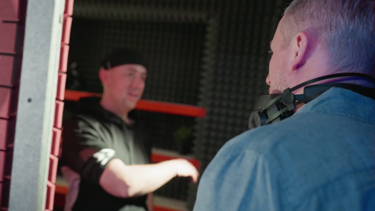 A focused dialogue between two men in a recording studio: one in a black cap and shirt, and the other in a blue shirt with headphones around his neck, against a red and black backdrop