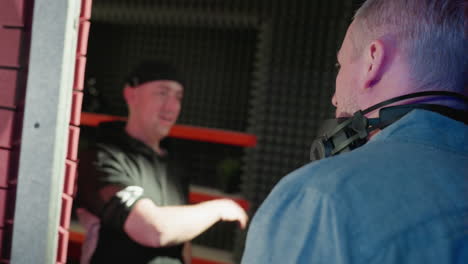 a focused dialogue between two men in a recording studio: one in a black cap and shirt, and the other in a blue shirt with headphones around his neck, against a red and black backdrop