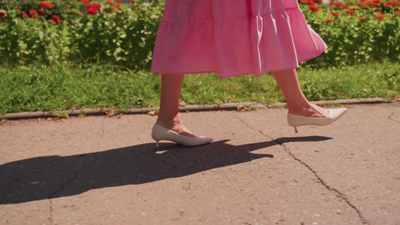 Abito rosa mentre cammina lungo un sentiero soleggiato, primo piano delle gambe e dei tacchi bianchi che proiettano una lunga ombra, leggero movimento della gonna, aiuole fiorite e prato verde sullo sfondo, ritmo nostalgico lento, ideale per la moda
