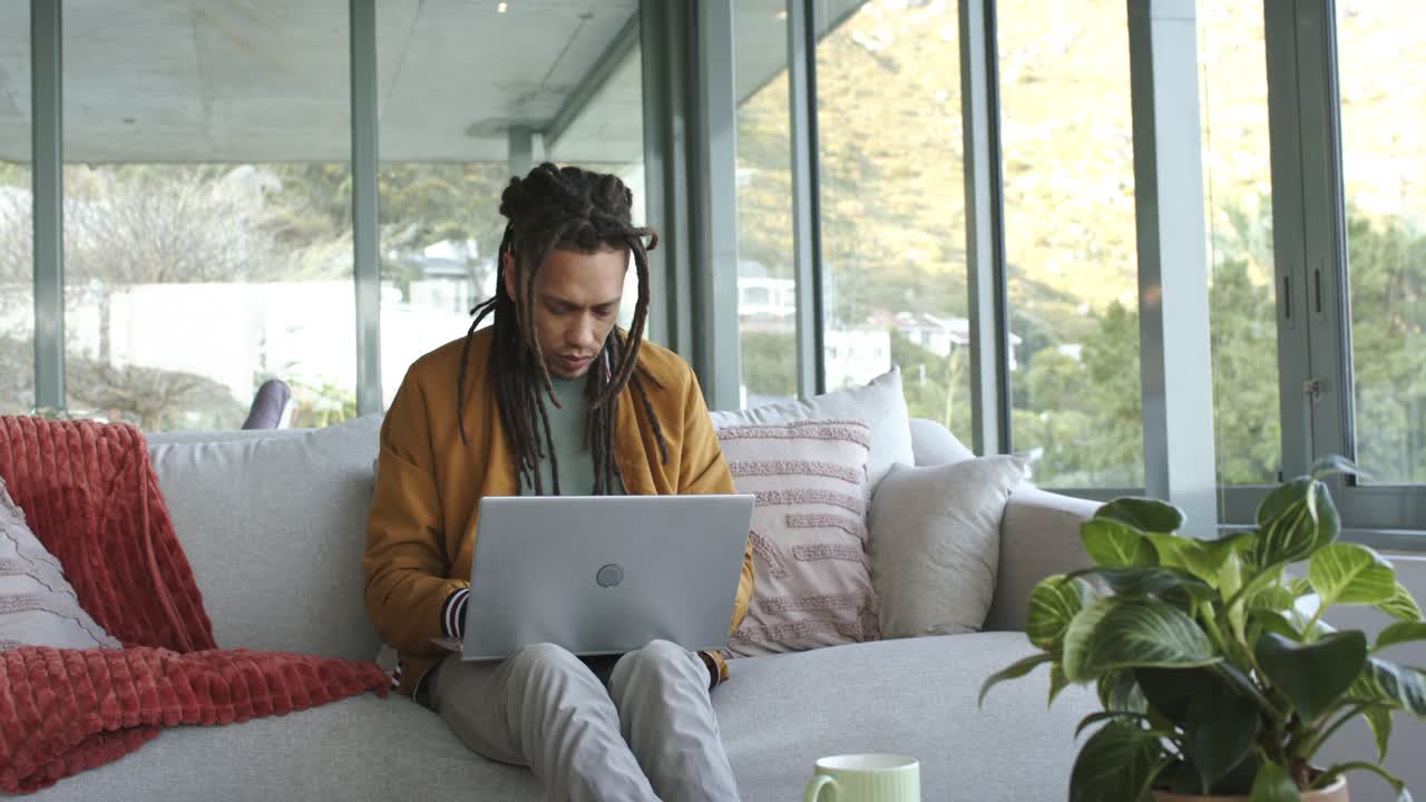 Mid adult African American man positioning laptop, typing, pausing to review work on sofa at home