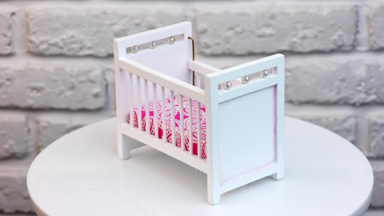 White bed for doll with decorations. Little white crib with bright pink mattress at the rotating stand. White wall background.
