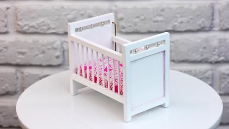 white bed for doll with decorations. little white crib with bright pink mattress at the rotating stand. white wall background.