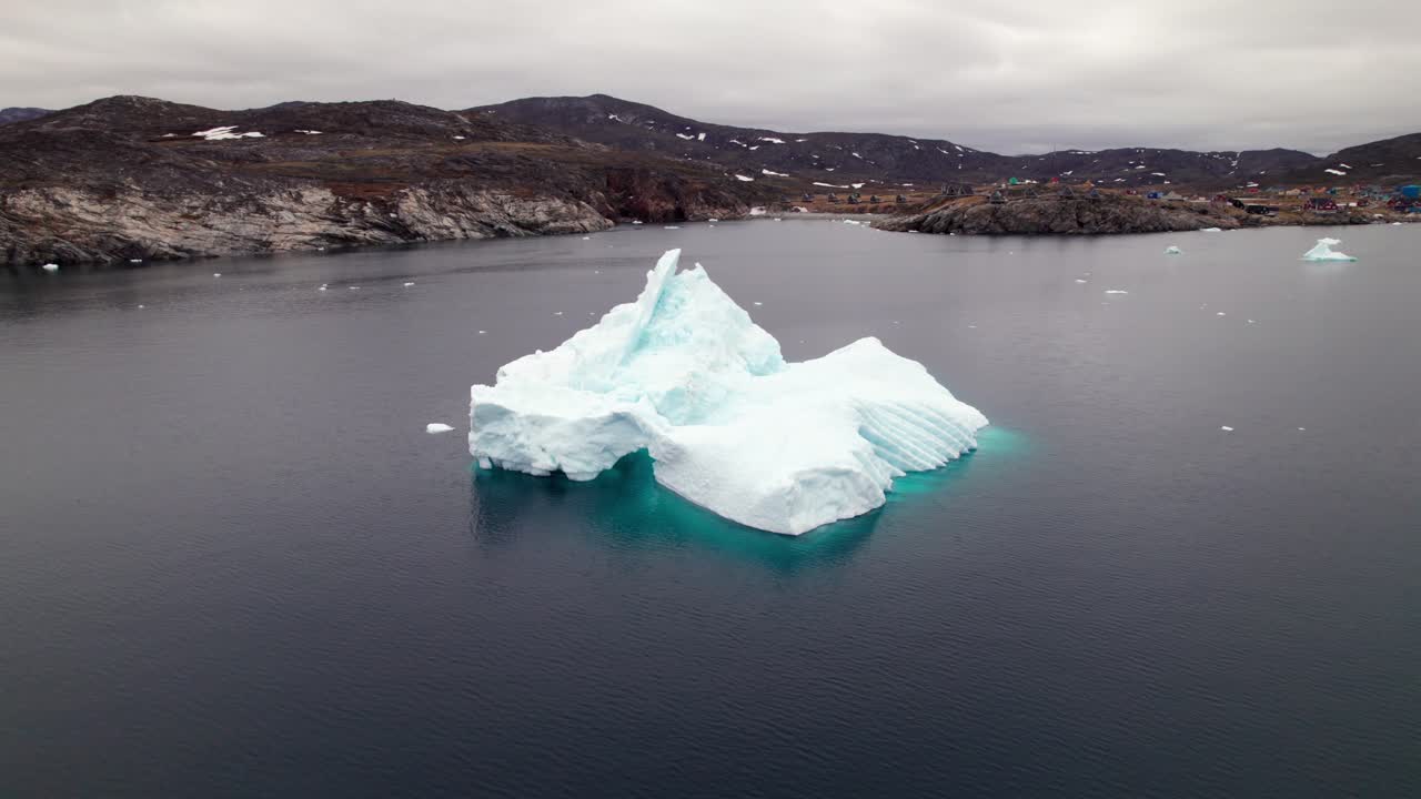 Circling drone shot around a massive iceberg in Greenland’s fjords near Ilimanaq, capturing the Arctic seascape and coastal settlement