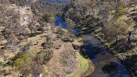 drone camera glides above a winding stream bordered by trees and grassy banks in ebor, new south wales, under bright natural daylight