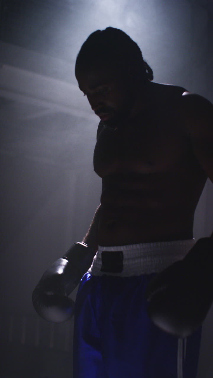 video de estudio vertical filmado con iluminación de baja tonalidad de boxeador silueta listo para luchar de pie en la piscina de luz en el ring de boxeo sparring