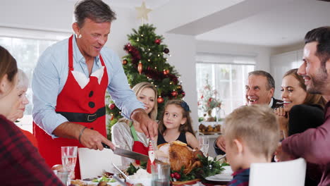 Un-Hombre-Tallando-Pavo-Mientras-Una-Familia-Multigeneracional-Se-Sienta-Alrededor-De-La-Mesa-Para-Una-Comida-Navideña-En-Casa-Juntos