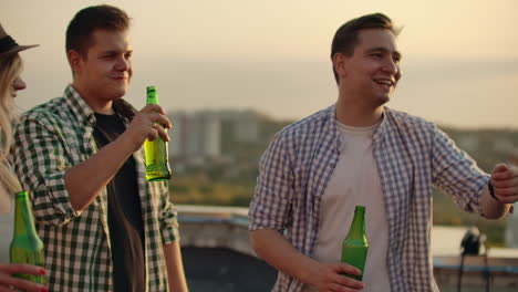 young men and women have enjoyable time on the roof. they smile and communicate with each other. they drink beer from green bottels and have fun in the plaid shirts.