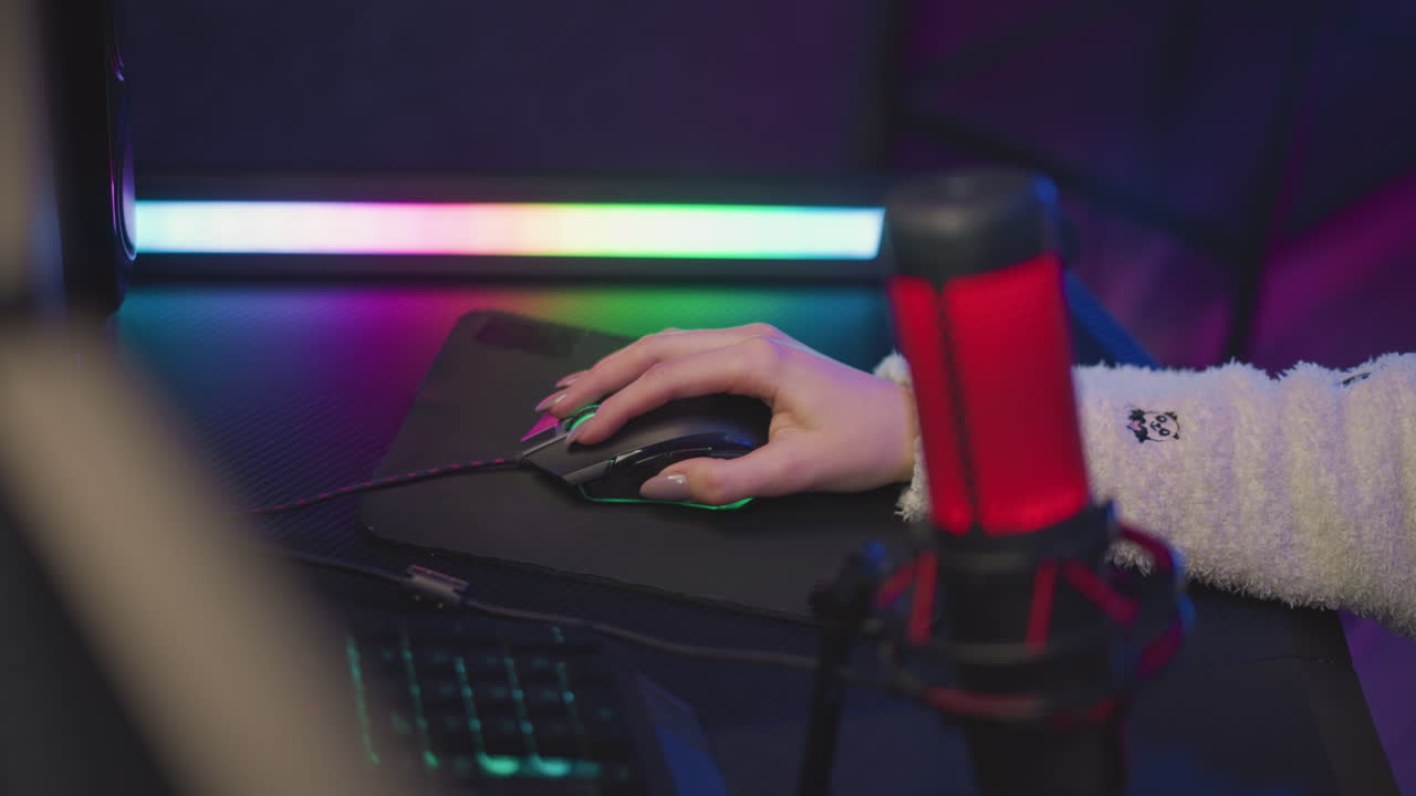 Close up lady hand with polished nail wearing fluffy sweater using wired gaming mouse on black pad, illuminated by soft pink and green ambient light