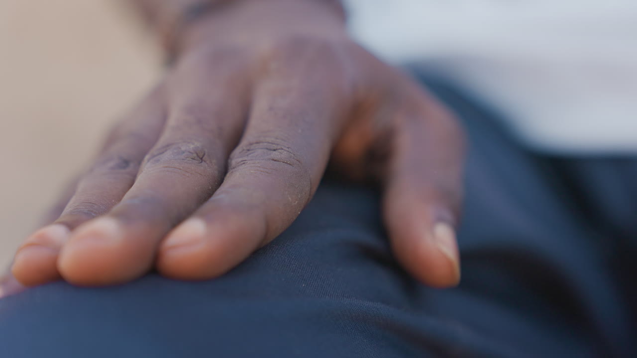 Nahaufnahme einer Handfläche eines schwarzen Läufers auf dem Oberschenkel. Ein schwarzer Sprinter stützt die Hand mit staubigen Fingerspitzen auf dunklen Trainingsshorts ab – ein angespannter Konzentrationsmoment im Freien vor dem Sprint, geprägt von Atemkontrolle und Entschlossenheit.