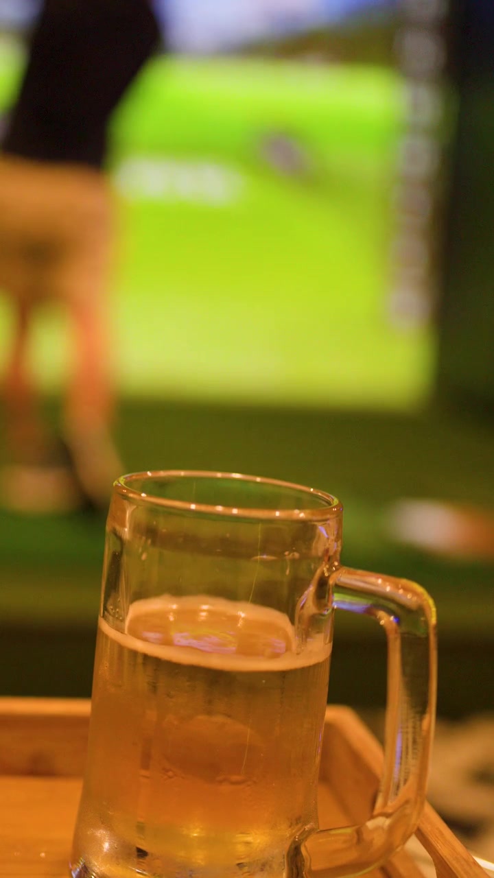 Una mano posa un boccale di birra fredda su un vassoio davanti a un simulatore di golf indoor.