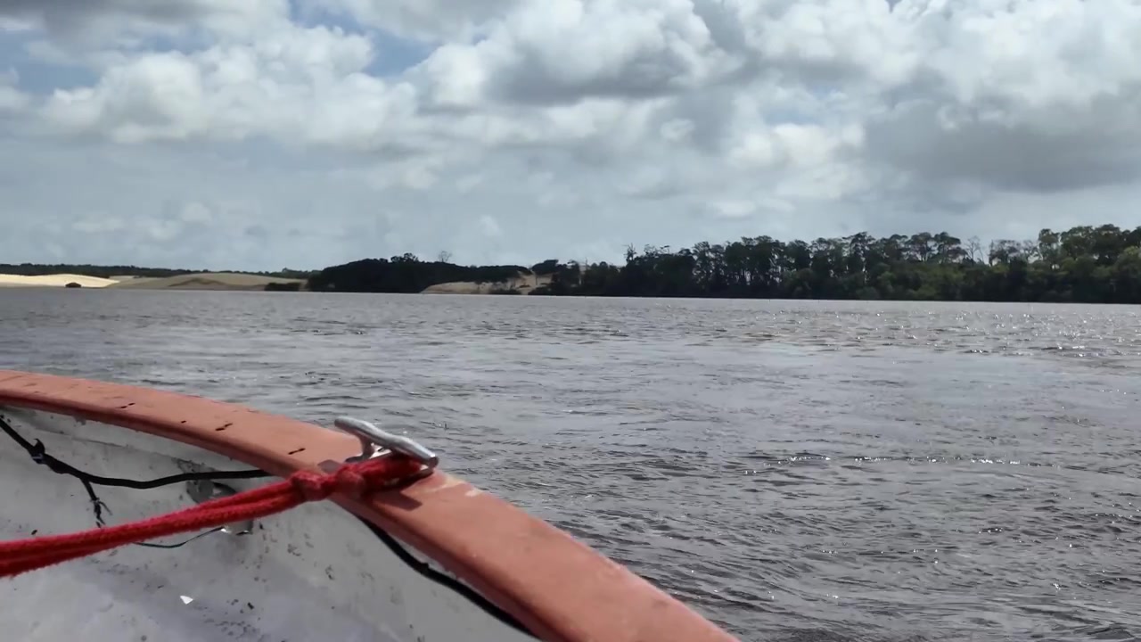 POV shot from the bow of a wooden boat cruising along the Preguiça River with lush green tree-lined shores under a cloudy sky in Maranhão, Brazil.