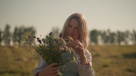 smiling gardener arranging fresh wildflowers under bright clear sky in picturesque farmland, woman wearing casual clothing, holding beautiful floral bouquet with distant blurred trees in background