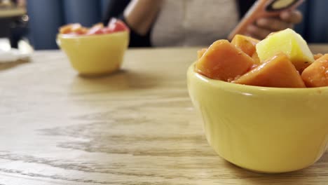 bowls filled with vibrant, fresh fruit salad sit on a restaurant table in mexico, while a customer enjoys a meal, savoring the flavors and browsing on a smartphone