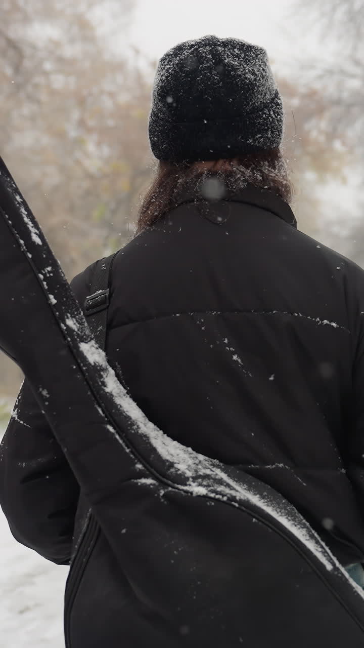 vista trasera de una persona caminando por un parque nevado con un paquete de guitarra en el hombro, rodeada de árboles helados y un paisaje invernal, con bancos y un camino de niebla que crea una atmósfera tranquila