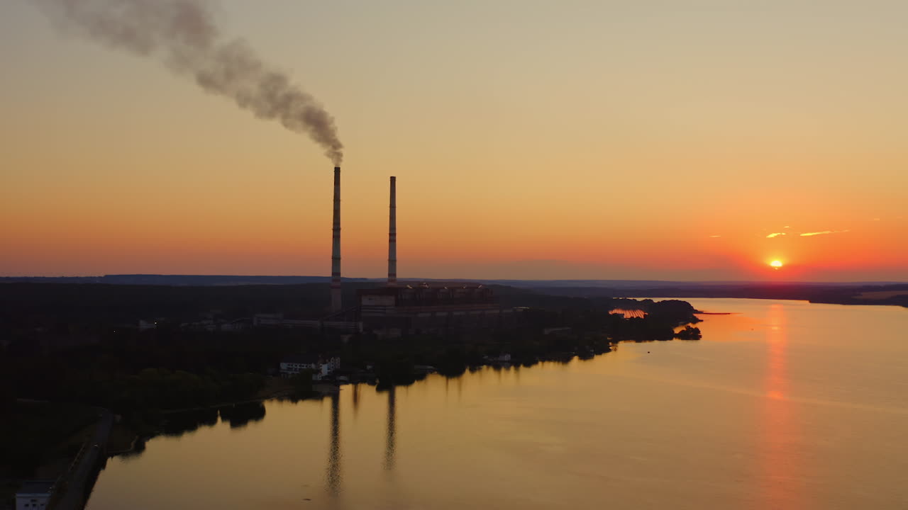 Factory releases smoke at sunset time with river side view. Dark silhouette of industrial plant with pipes with smoke in the evening. Ecology suffers from harmful gases.