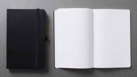 Nahaufnahme-Eines-Offenen-Leeren-Buches-Und-Eines-Schwarzen-Notizbuchs-Mit-Kopierraum-Auf-Grauem-Hintergrund-In-Zeitlupe