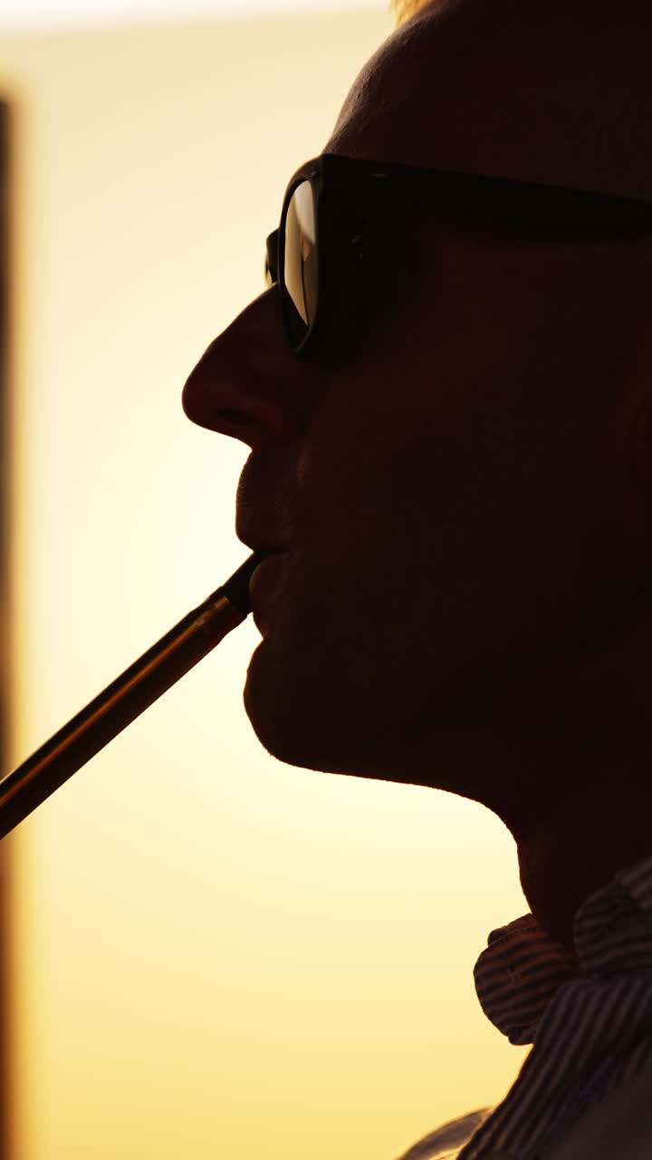 Upper part of a man in sunglasses breathing in and out thick white smoke. Young man smoking hookah at sunset close up. Vertical video