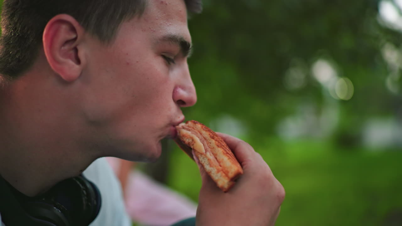 Close-up of man eating grilled sandwich outdoors, slightly blurred background of another person eating, set in a natural environment with soft lighting