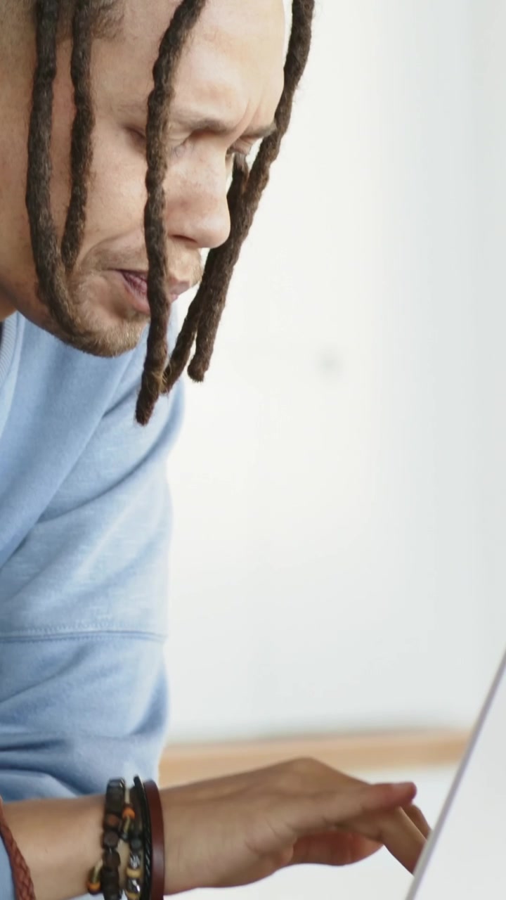 Vertical video: Pulling back man in blue top with bracelets typing on laptop at desk, copy space