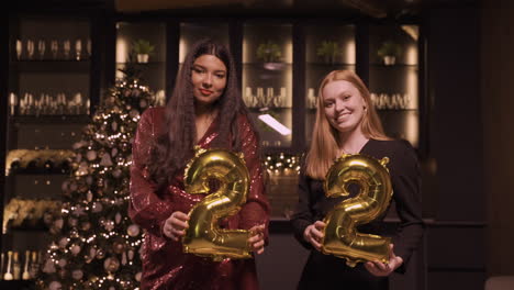 two women friends wearing elegant clothes holding balloons with the numbers 22 while dancing in new year's party