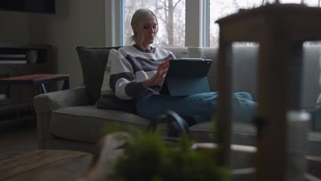 woman sitting on the couch in front of a bay window, drinking coffee while scrolling on a tablet with her feet up on the couch from a diagonal angle