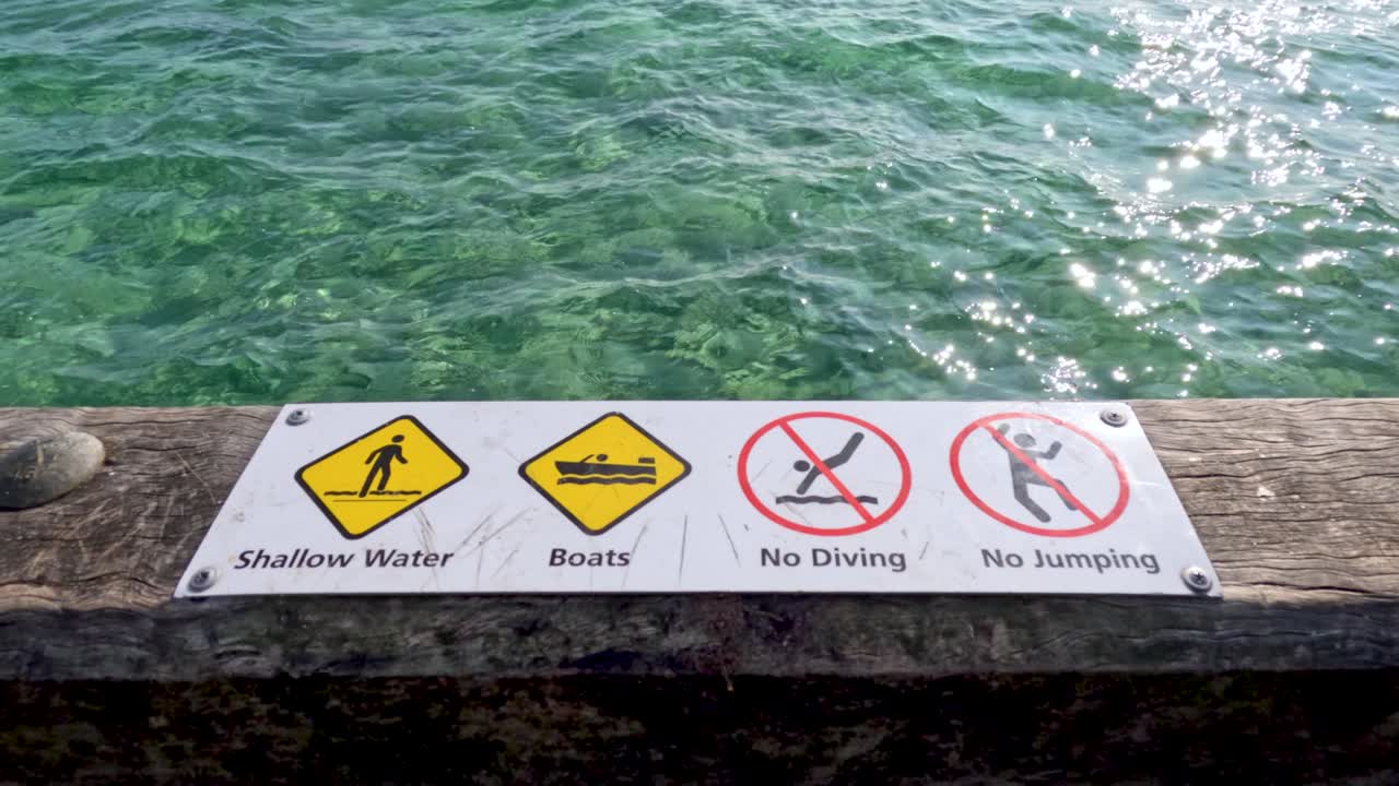 A series of safety signs on a pier warn against diving, jumping, and boats in shallow waters