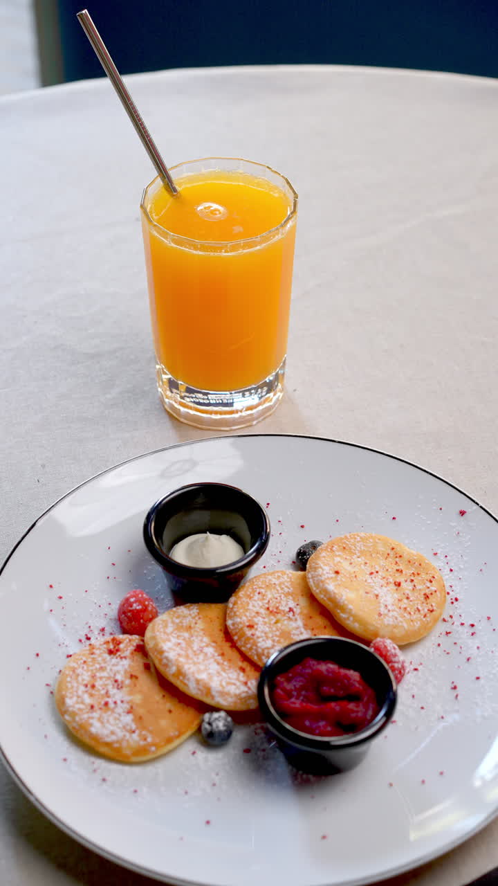 Homemade breakfast pancakes adorned with fresh berries, jam, and cream, accompanied by orange juice, elegantly arranged on beige round table surface