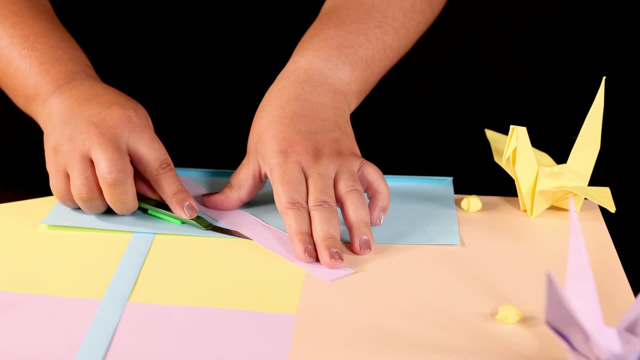 Child’s hands fold pastel paper for origami, surrounded by cranes, under bright studio lighting