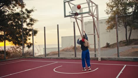 a blonde girl in white pants throws a basketball to her friend who throws it into the basket on a red-colored basketball field early in the morning at sunrise