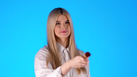 Modelo-Femenino-Blanco-Atrapa-Un-Cepillo-Cosmético-Y-Aplica-Maquillaje,-Fondo-Azul