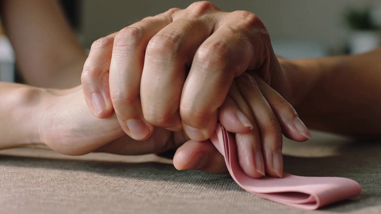 Mão adulta maior envolvendo mão adulta menor em mesa doméstica, puxando pano rosa para conforto
