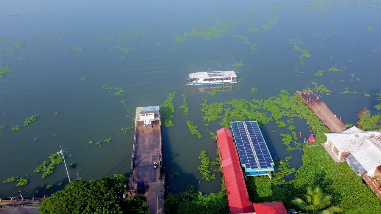 Aerial view of a solar-powered passenger ferry moving away from a dock, surrounded by lush green aquatic plants and traditional waterfront buildings in Kerala, India