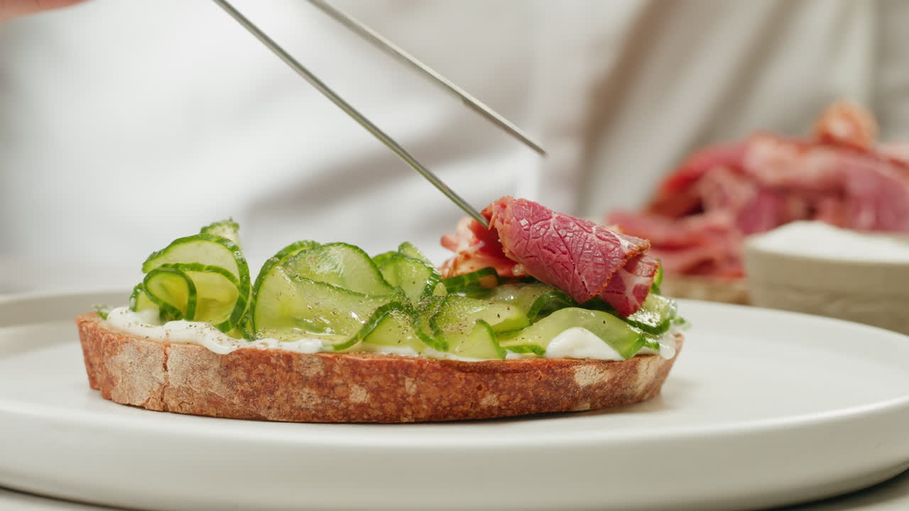Chef preparing a gourmet sandwich with sliced cured meats and cucumbers