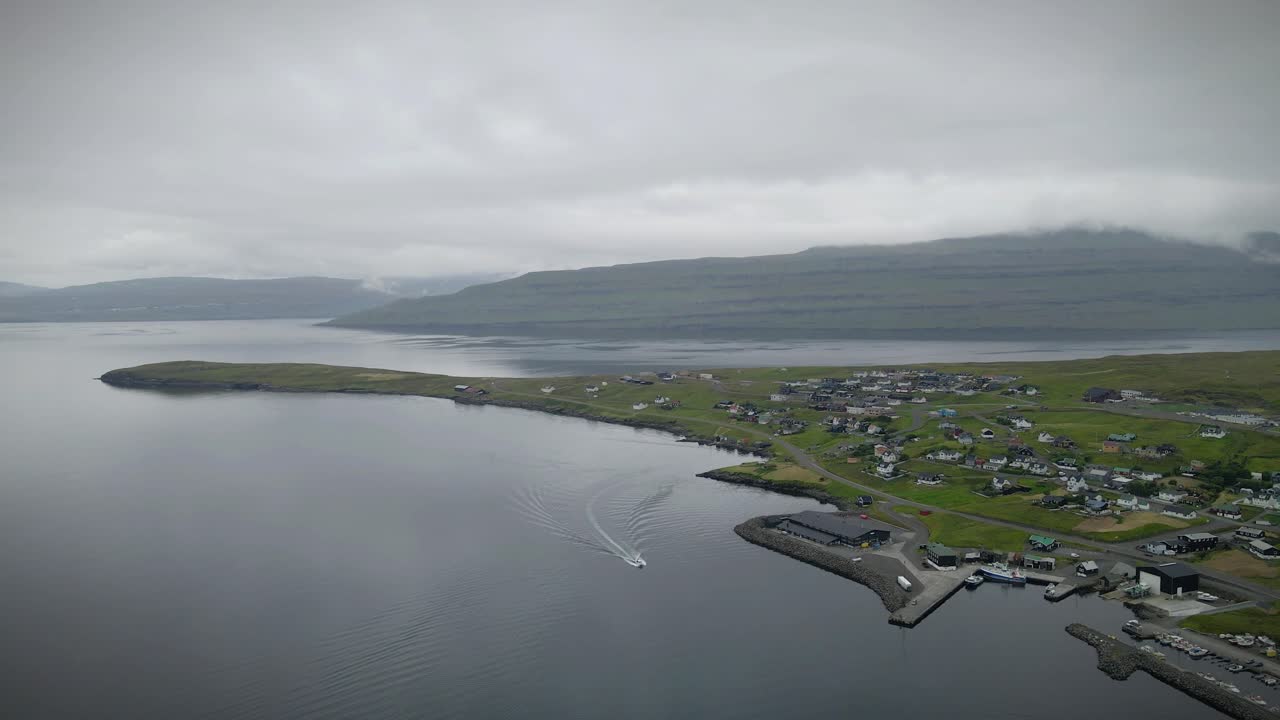 드론은 파로 제도의 스트렌두르 (strendur) 해안가를 따라 우아하게 항해하는 배를 포착합니다. 구름이 가득한 하늘 아래, 잔잔한 녹색 언덕이 배경으로 아 있습니다.