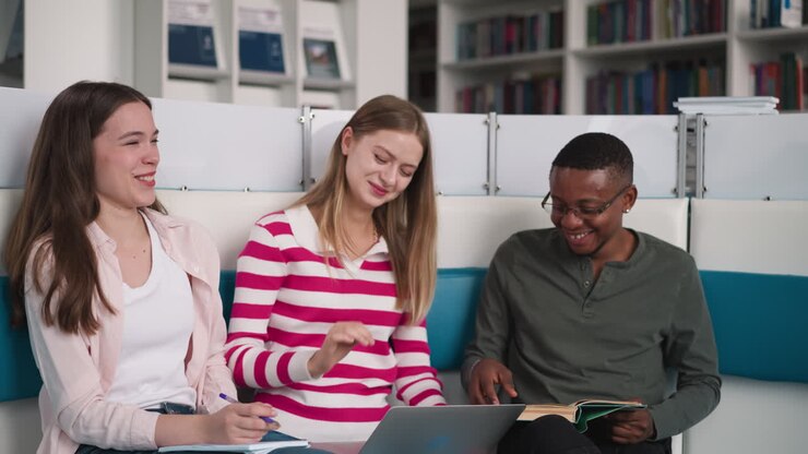 Friends of different races with laptop laugh in library. Happy students team has fun preparing educative project materials in reading hall. E-learning integration