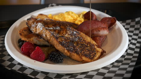 Maple-syrup-drizzles-over-french-toast-dusted-with-powdered-sugar-paired-with-fresh-berries-bacon-and-scrambled-eggs,-slow-motion-close-up-slider-4K