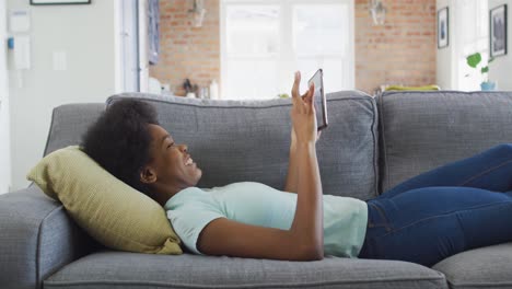 Happy-african-american-teenage-girl-lying-on-sofa,-using-tablet